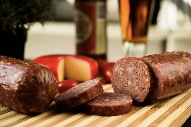 Summer sausage log and sliced rounds on a cutting board with cheese and beer